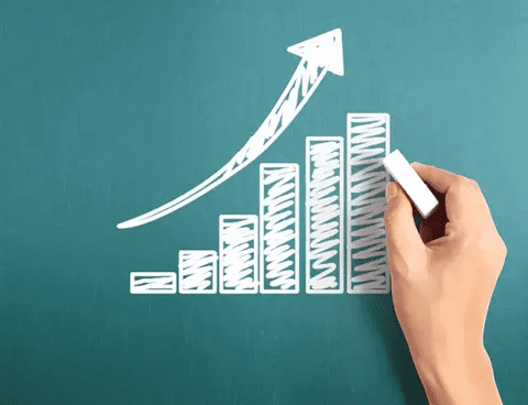 Hand drawing a rising bar chart with an upward arrow on a green board.